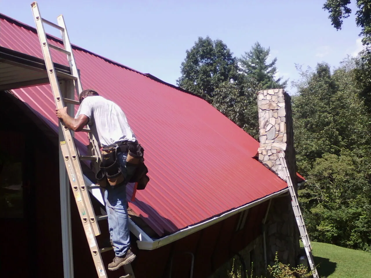 Roofer on a ladder beside a red metal roof and stone chimney — Soffit & Fascia Repair by Roofing Brothers in Lansdowne